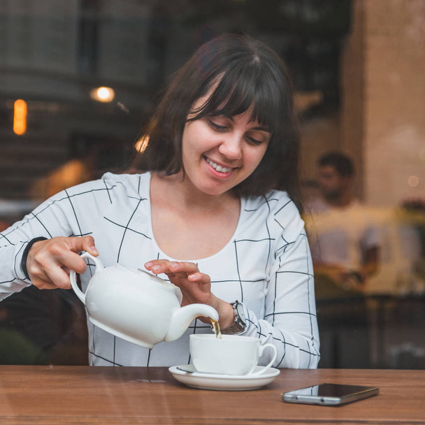 женщина наливает чай в чашку в кафе. вид через стеклянное окно
 - Фото, изображение