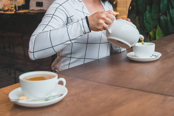 Frau gießt Tee aus weißem Wasserkocher auf Mütze im Café. Aufwärmen - Foto, Bild