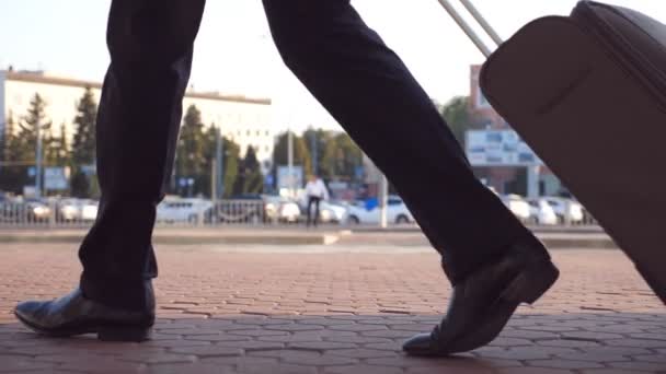 Nohy úspěšný podnikatel chůzi na ulici města a táhne kufr na kolečkách. Nohy z mladých mužů podnikání osoby, které bude s jeho zavazadly na městské prostředí po letu. Slow motion boční pohled - Záběry, video
