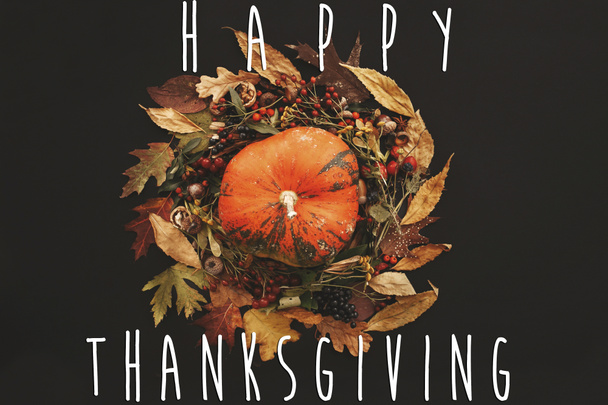 Happy Thanksgiving text on pumpkin in Fall leaves wreath with berries, nuts, acorns, flowers,herbs on black background. Autumn composition. Seasons greetings card. - Photo, Image