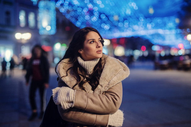 Schattig jong meisje in winterjas permanent in straat en genieten van prachtige winteravond in versierde straten. - Foto, afbeelding