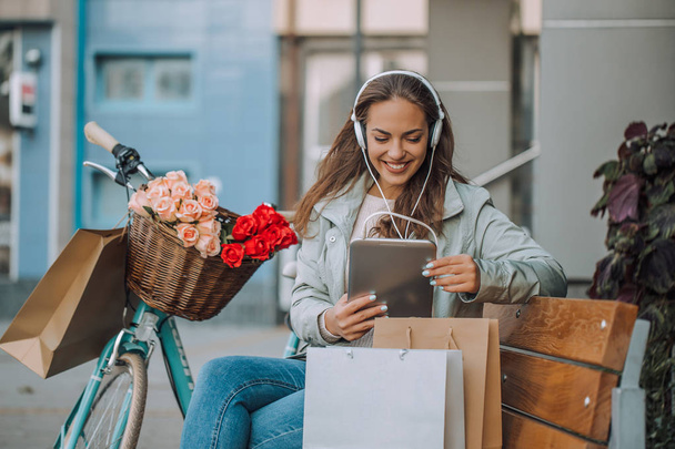 Mujer joven y feliz escuchando música a través de auriculares y tabletas digitales mientras está sentada en el banco de la ciudad
. - Foto, imagen
