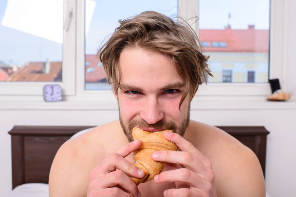Guy holds croissant bedroom hotel room window background. Enjoy every bite. Man eats croissant he likes bakery products. Guy bites sweet dessert. Man bearded handsome guy eating breakfast in bed - Photo, Image