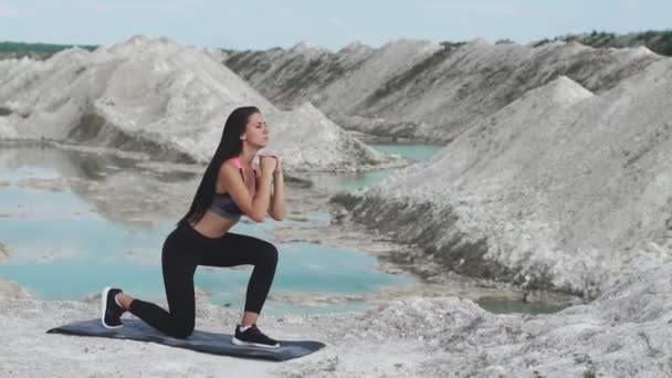 Sporty brunette girl in black sportswear trains against a white chalk sand quarry with blue water. Lunges on each leg for thighs - Footage, Video