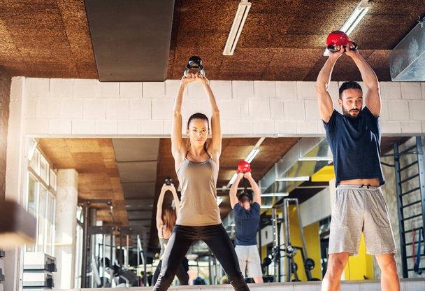 Strong young happy couple lifting kettle bells in a gym. Standing in front of a camera. - Фото, изображение