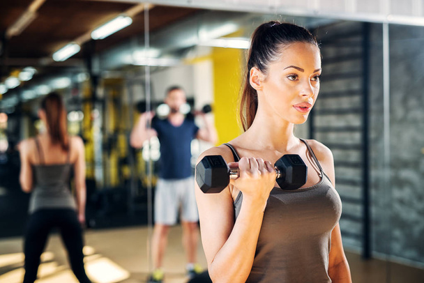 Portrait of a beautiful focused young sporty female in a gym holding weight in one hand, - Photo, Image