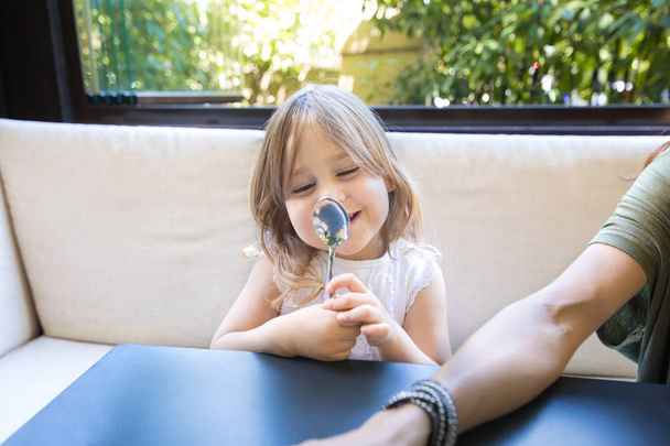 retrato divertido de niña rubia de cuatro años, mirándose en una cuchara y haciendo una cara y sonriendo, sentada en un restaurante
 - Foto, Imagen