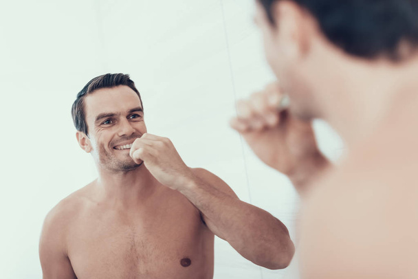 Mirror Reflection of Man Brush Teeth in Bathroom. Portrait of Smiling Handsome Brown Haired Young Person with Bare Chest Holding Toothbrush and Looking at Mirror. Morning Concept - Foto, Imagem