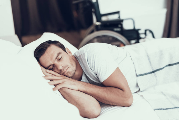 Person Sleeps Near Alarm in Bed With White Linens. Handsome Young Man Lying in Bedroom with Blurred Wheelchair on Backround Keeping Hands Under Head and Wearing White T-shirt. Morning Concept - 写真・画像