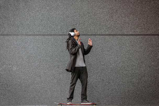 Jonge stedelijke dreadlocks hipster man rijden op zijn lange bord en luisteren naar de muziek. Rijden op straat met de zwarte muur in achtergrond. - Foto, afbeelding
