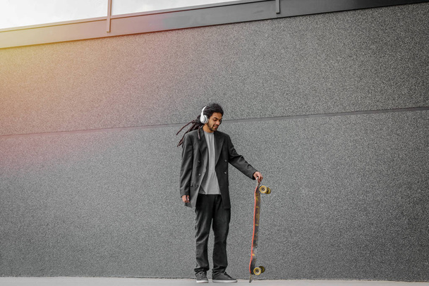 Moderno urbano jovem dreadlocks hipster homem segurando seu skate na rua e ouvir música. De pé contra a parede preta com fones de ouvido na cabeça
. - Foto, Imagem