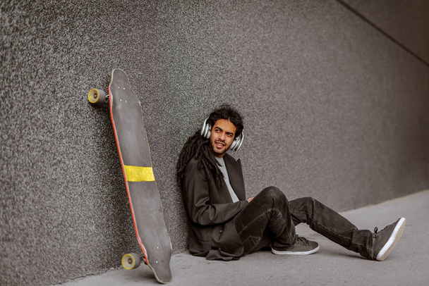 Jovem hipster dreadlocks homem sentado na rua inclinado na parede preta e ouvir a música, enquanto seu skate é inclinado na parede. Relaxar e se divertir
. - Foto, Imagem