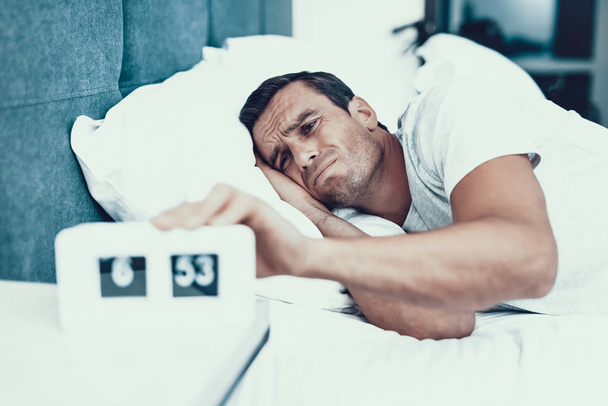 Man Hardly Awakes and Pulls Alarm Off in Morning. Handsome Young Person in Bad Mood Lying in Bedroom in Bed With White Linens Near Alarm Clock and Wearing White T-shirt. Morning Concept - Φωτογραφία, εικόνα