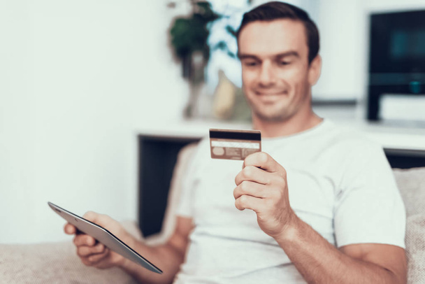 Blurred Person Holds Credit Card and Uses Tablet. Handsome Smiling Man Sitting on Couch Does Online Shopping and Showing Satisfied Face Expression in Modern Living Room at Home - Photo, Image