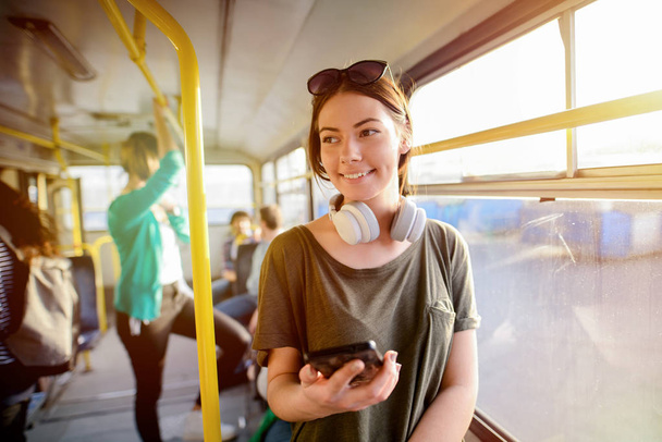 Современная улыбающаяся милая девушка, стоящая в автобусе с наушниками на шее. Улыбаясь и отворачиваясь
. - Фото, изображение