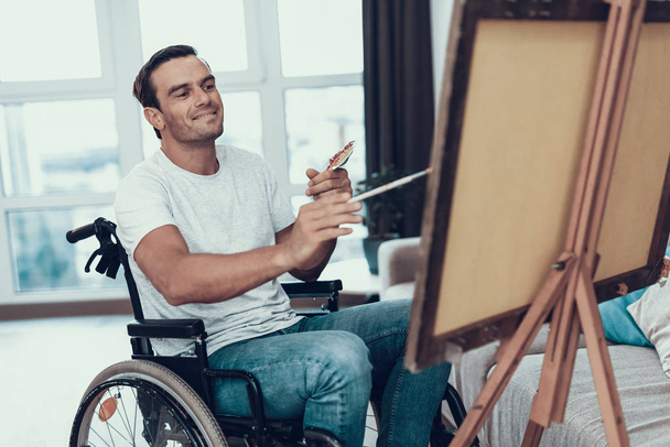 Disabled Man in Wheelchair Painting Picture. Smiling Handsome Happy Caucasian Person Sitting Opposite Easel in Living Room Wearing Casual Clothes Holding Paintbrush and Palette - Photo, image