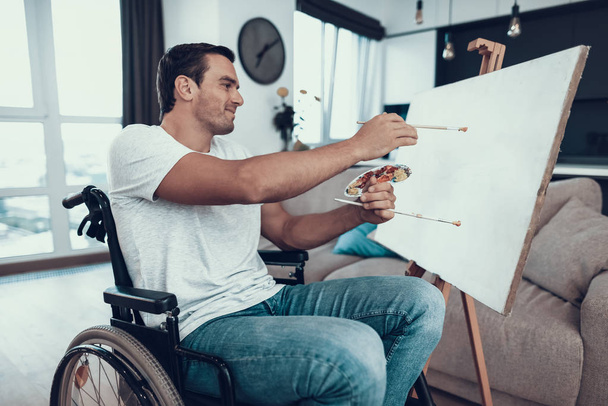 Handsome Disabled Young Man Painting Picture. Portrait of Smiling Caucasian Person in Wheelchair Opposite Easel Wearing Casual Clothes Holding Paintbrushes While Sitting in Living Room at Home - Foto, Imagem