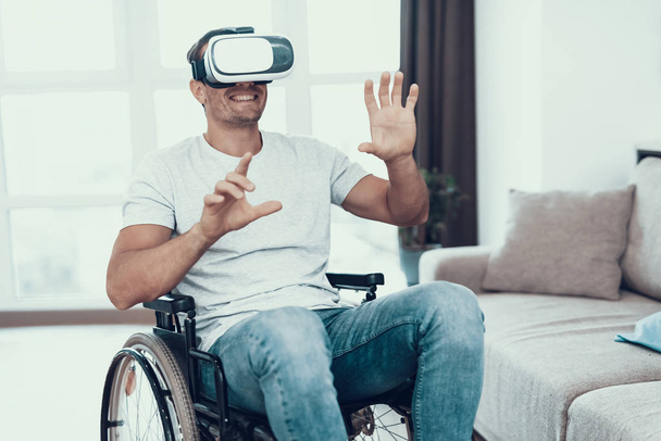 Disabled Man with Hands Up Wearing VR Goggles. Portrait of Smiling Person Sitting in Wheelchair Parked in Large Bright Living Room Wearing T-Shirt and Blue Jeans Enjoying Virtual Reality Headset - Fotografie, Obrázek