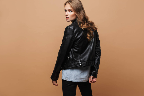 Young beautiful woman with wavy hair in black leather jacket thoughtfully looking in camera from back over beige background - Photo, Image