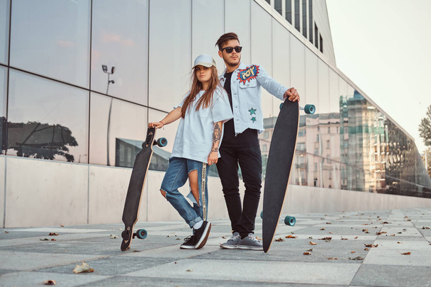 Attractive young couple dressed in trendy clothes posing with skateboards near skyscraper. - Valokuva, kuva