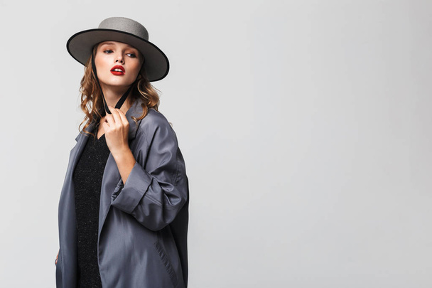 Young beautiful woman with wavy hair in modern hat and cloak thoughtfully looking aside over gray background - Photo, Image