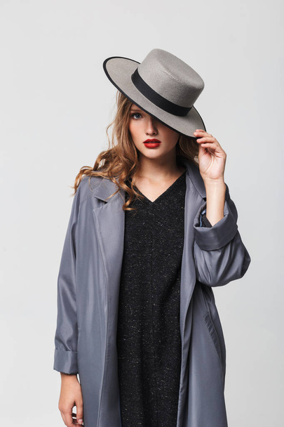 junge attraktive Frau mit welligem Haar im Mantel, die Augen mit Hut bedeckt und nachdenklich in die Kamera über grauem Hintergrund blickt - Foto, Bild