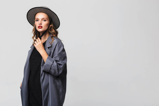 junge attraktive Frau mit welligem Haar in modernem Hut und Mantel, die nachdenklich in die Kamera über grauem Hintergrund blickt - Foto, Bild