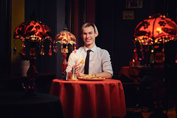Красивый молодой человек в белой рубашке и галстуке ест начос в мексиканском ресторане
. - Фото, изображение