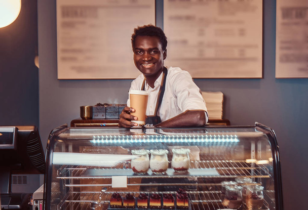 Portret van een knappe Afrikaanse barista in wit overhemd en schort houdt kop van koffie en ontspannen na een werkdag bij koffie shop. - Foto, afbeelding