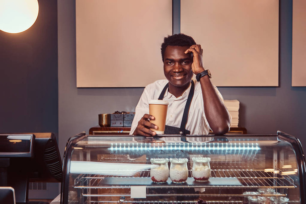 Muotokuva komea Afrikkalainen barista valkoinen paita ja esiliina pitää kupin kahvia ja rentouttava työpäivän jälkeen kahvilassa
. - Valokuva, kuva