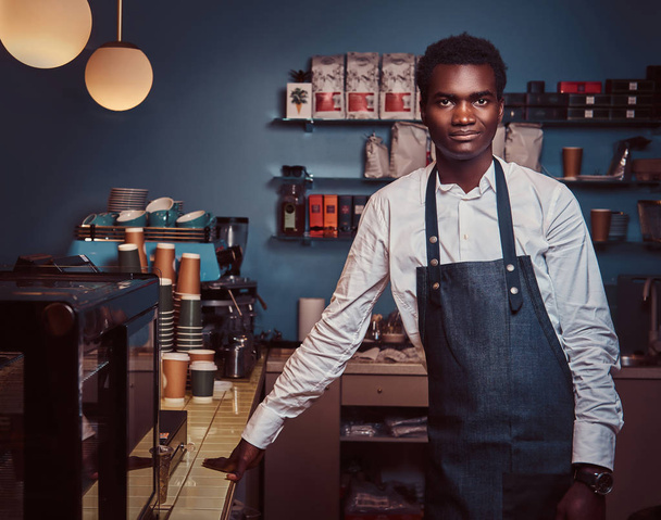 Αφρικανική αμερικανική barista χαμογελά στη φωτογραφική μηχανή και να στέκεται welcomingly στο μαγαζί καφέ της. - Φωτογραφία, εικόνα