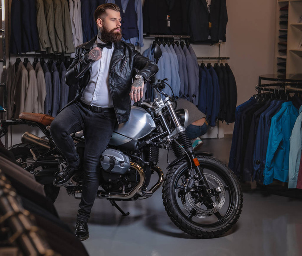 Homem barbudo tatuado elegante com vestido de jaqueta de couro preto e gravata borboleta posando perto de moto retro esportes na loja de roupas dos homens
. - Foto, Imagem