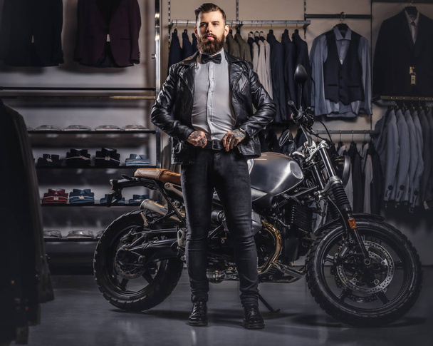 Elegante hombre barbudo tatuado con chaqueta de cuero negro y pajarita posando cerca de la moto deportiva retro en la tienda de ropa para hombre
. - Foto, Imagen