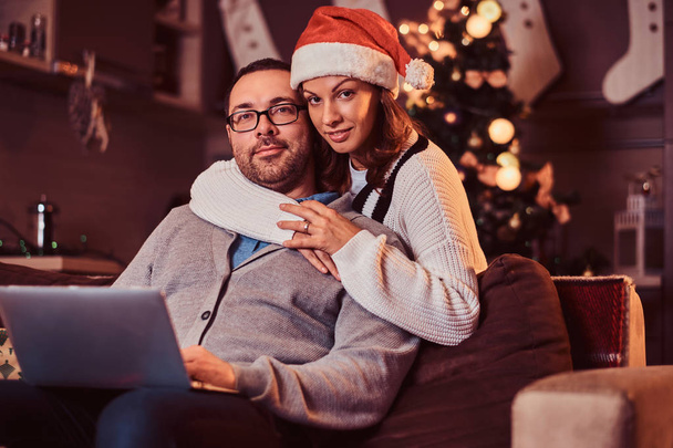 Portrait of an adorable couple with laptop - charming woman in Santa hat hugging her man and looking at camera. - Photo, Image