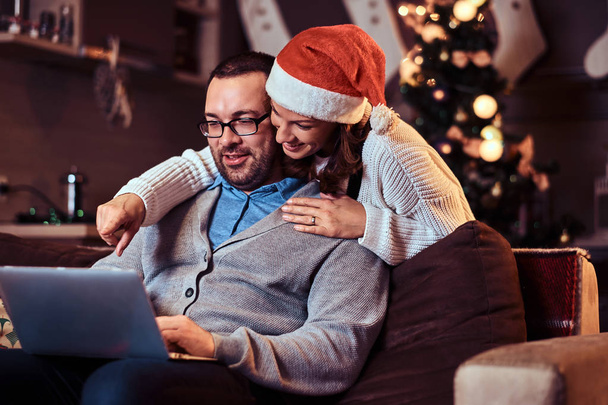 Portret urocza para - uroczy Kobieta w Santa hat przytulanie mężczyznę i za pomocą laptopa. - Zdjęcie, obraz