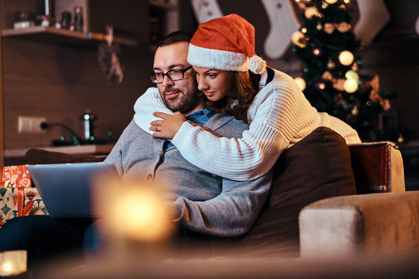 Portrait d'un couple adorable - charmante femme dans le chapeau du Père Noël embrassant son homme et utilisant un ordinateur portable
. - Photo, image