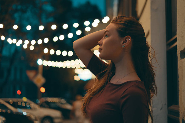 Halve lengte portret van een aantrekkelijk meisje gekleed in casual kleding met nachtverlichting op de achtergrond. Romantische vrouwelijke staande in de straat verlichting. - Foto, afbeelding