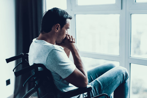 Disabled Person in Wheelchair Looking at Window. Closeup of Sad Serious Caucasian Man Wears White T-Shirt and Jeans, Keeps Hands near Mouth and Shows Thoughtful Face Expression. - Foto, Bild