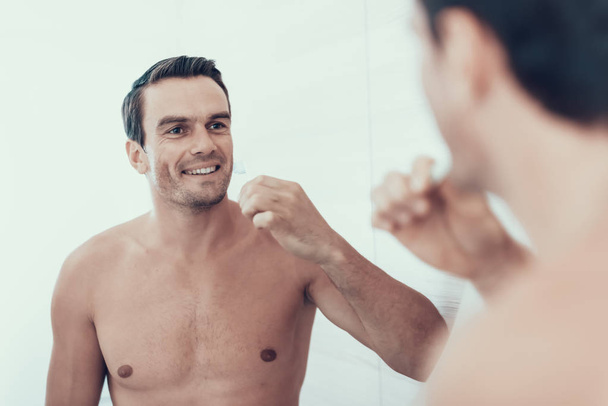 Mirror Reflection of Man Brush Teeth in Bathroom. Portrait of Smiling Handsome Brown Haired Young Person with Bare Chest Holding Toothbrush and Looking at Mirror. Morning Concept - Foto, immagini