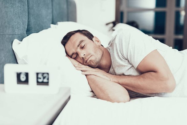 Person Sleeps Near Alarm in Bed With White Linens. Handsome Young Man Lying in Bedroom in Modern Apartment Keeping Hands Under Head and Wearing White T-shirt. Morning Concept - Photo, Image