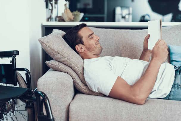 Man Lying on Couch with Tablet near Wheelchair. Cheerful Handsome Young Smiling Disabled Person Relaxing on Comfortable Bright Couch in Modern Apartmentand Surfing in Internet - Φωτογραφία, εικόνα