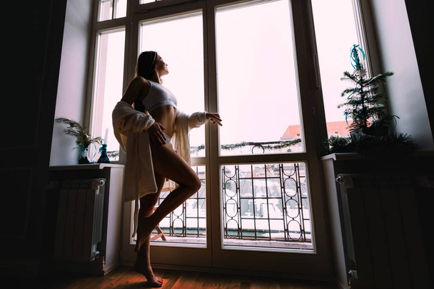 Hermosa chica sexy en bata blanca de pie cerca de la ventana con en apartamento moderno de lujo, mirando paisaje urbano después de despertar por la mañana, relajante. Mañana perfecta
. - Foto, Imagen