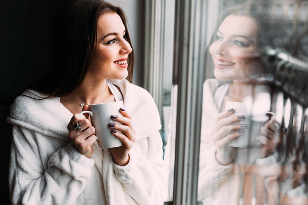 Splendida ragazza sognante sorridente in vestaglia bianca stand vicino alla finestra con una tazza di caffè o tè. Caffè del mattino, concetto di felicità
. - Foto, immagini