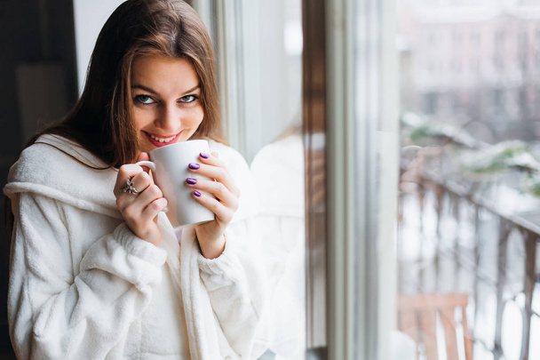 Όμορφο κορίτσι στέκεται δίπλα στο παράθυρο. Είναι χαμογελαστός και κρατώντας ένα φλιτζάνι καφέ, τσάι στο χέρι. Περιβάλλεται από τον ήπιο πρωινό ήλιο που λάμπει μέσα από το παράθυρο γυαλιού. - Φωτογραφία, εικόνα
