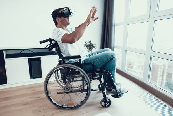 Handicapped Man in Wheelchair Wearing VR Headset. Portrait of Male Disabled Person in Casual Clothes Sitting Near Window in Large Bright Room and Enjoying Virtual Reality Glasses and Keeping Hands Up - Foto, Imagem