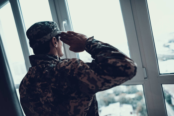 Ernst in Militäruniform salutiert am Fenster. Rückansicht eines selbstbewussten kaukasischen Mannes mit Hut, der die Hand in der Nähe des Kopfes in einer modernen Wohnzimmerwohnung hält. Soldatenkonzept - Foto, Bild