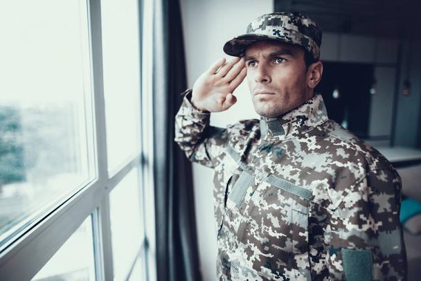 Portret van militaire Uniform Salute in de buurt van venster. Zelfverzekerde Kaukasische Man met hoed houdt Hand in de buurt van hoofd permanent in moderne woonkamer appartement en ernstige gezicht expressie te tonen - Foto, afbeelding