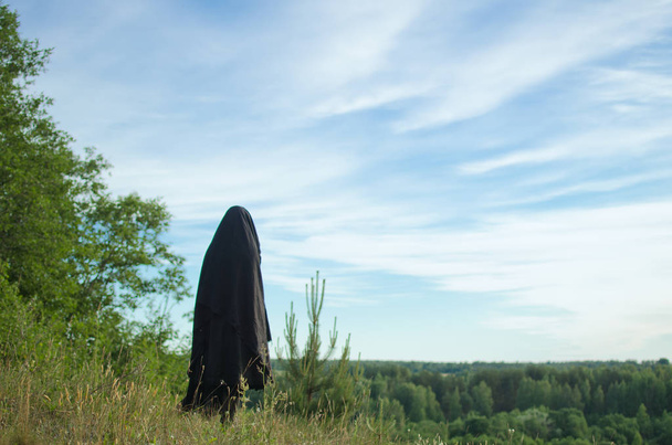 黒マントの女魔女が草で覆われた山から森の中に見える - 写真・画像