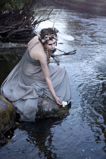 Młoda kobieta siedzi na skale w pobliżu rzeki i pozwala float biały kwiat do wody - Zdjęcie, obraz