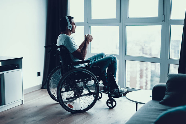 Инвалид слушает музыку и пьет кофе. Крупный план Грустный европеец в современном инвалидном кресле носит наушники и смотрит на большое панорамное окно в гостиной или больнице
 - Фото, изображение